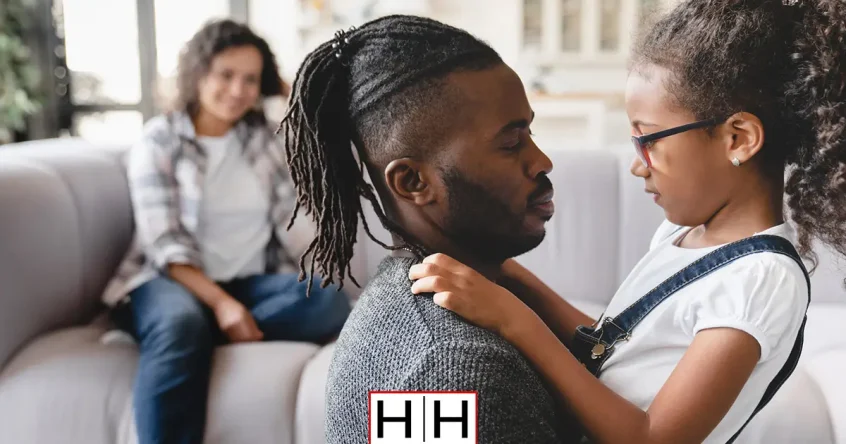 A father holds his young daughter on a couch while a woman sits in the background, illustrating a family affected by a fault vs. no-fault divorce case in Salt Lake City, Utah.